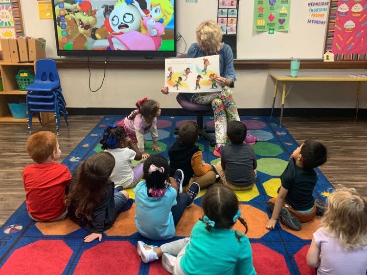 Class sitting on the floor while teacher reads a book towards class